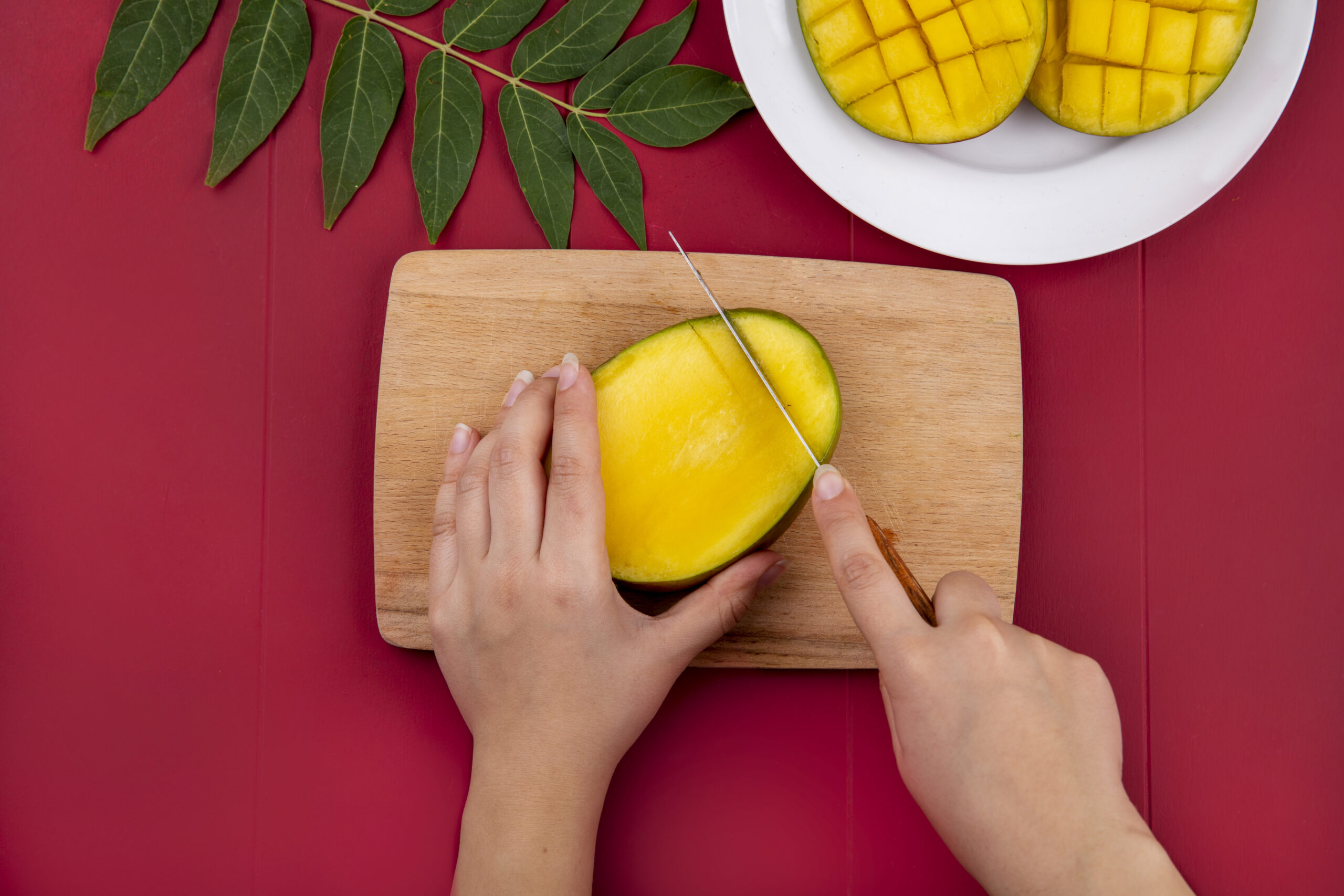 top view of female hands cutting mango with knife on woodenkitchen board with sliced mango in a white plate and with green leaf on red background morocco